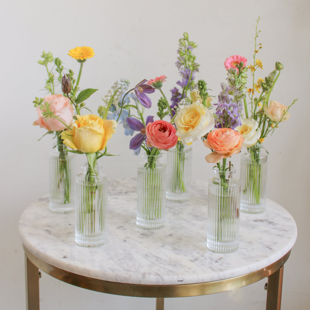 A set of clear glass bud vases with on a marble table in front of a white backdrop. They're filled with pink, peach, yellow blue and light purple flowers, including rose, piccolini daisy, clematis, ranunculus, and delphinium.