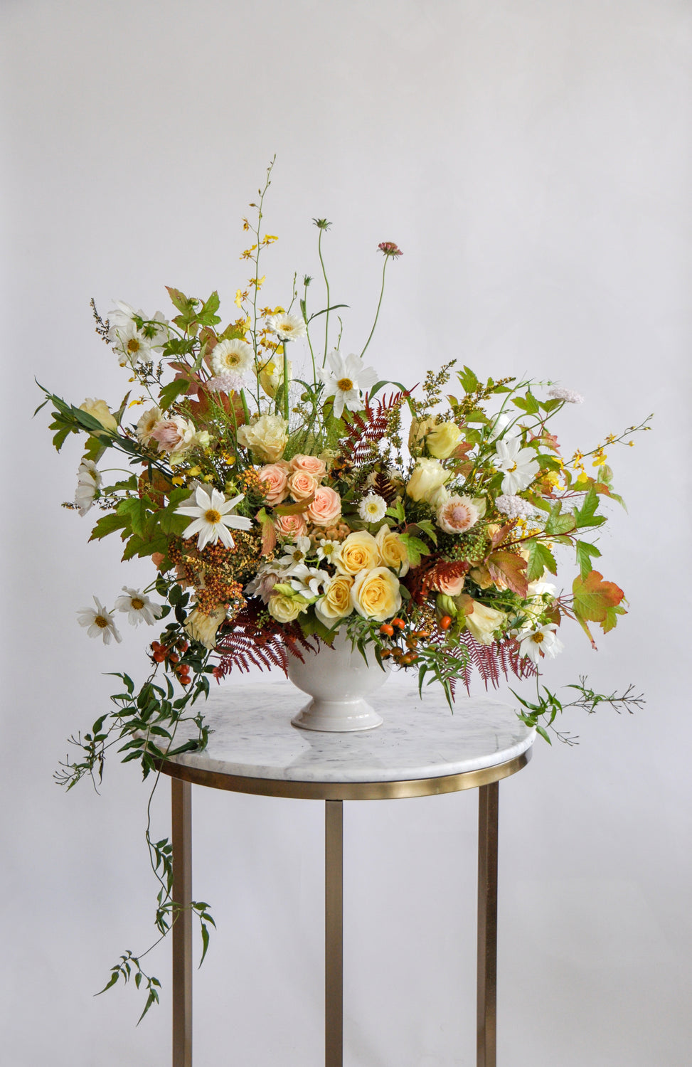 A green, yellow, peach, and maroon WildFlora flower arrangement in a white vase, featuring rose, lisianthus, fern, and oncidium orchid, cosmos, fall foliage leaves, protea, gerber daisy, and berries.