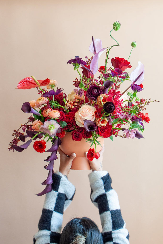 A woman in a checkered sweater holds a large flower arrangement in a terracotta container in front of a tan backdrop. It's filled with red, purple, peach, maroon, and dark violet-colored flowers, including rose, ranunculus, orchid, calla lily, anthurium, hellebore, and allium.