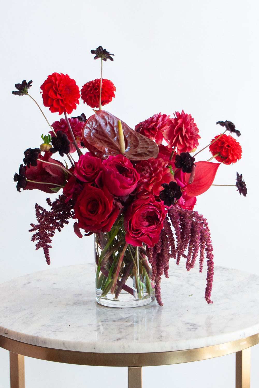 A floral arrangement featuring a variety of red and black flowers, placed in a clear glass vase on a marble table with a gold frame.