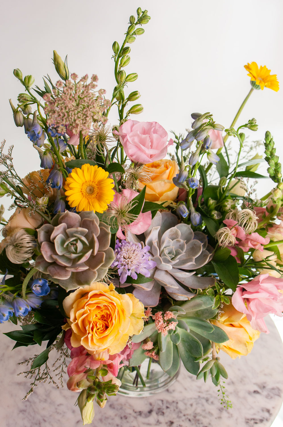 A flower arrangement in a glass vase in front of a white backdrop on a small marble table. It's filled with pastel pink, yellow, green, blue and purple flowers, including succulents, rose, delphinium, piccolini daisy, lisianthus, scabiosa, Queen Anne's lace, and tulip.
