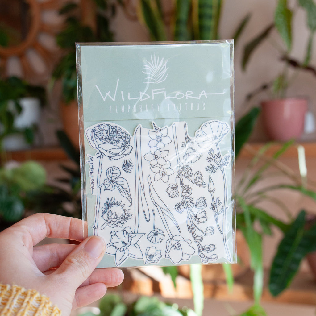 Hand holding WildFlora temporary tattoos with floral designs against a plant-filled background