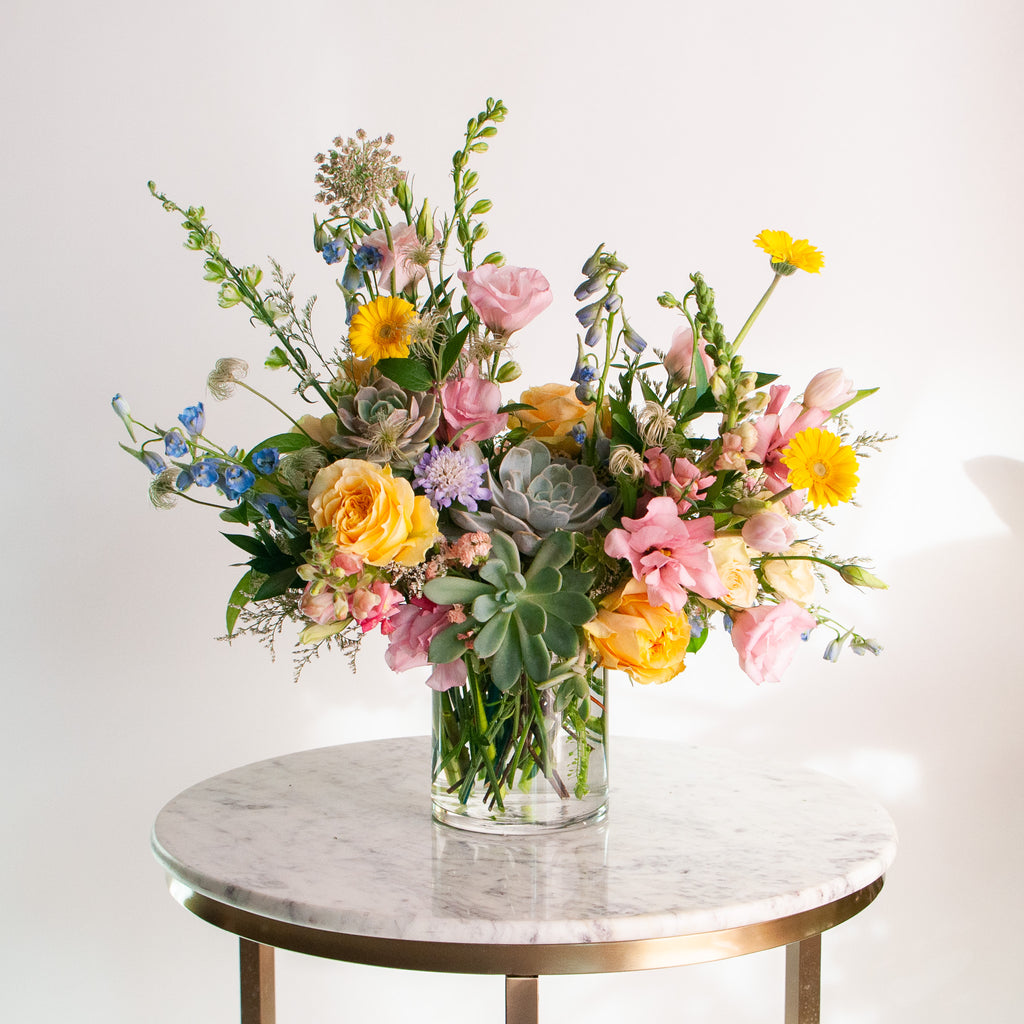 A flower arrangement in a glass vase in front of a white backdrop on a small table. It's filled with pastel pink, yellow, green, blue and purple flowers, including succulents, rose, delphinium, piccolini daisy, lisianthus, scabiosa, Queen Anne's lace, and tulip.