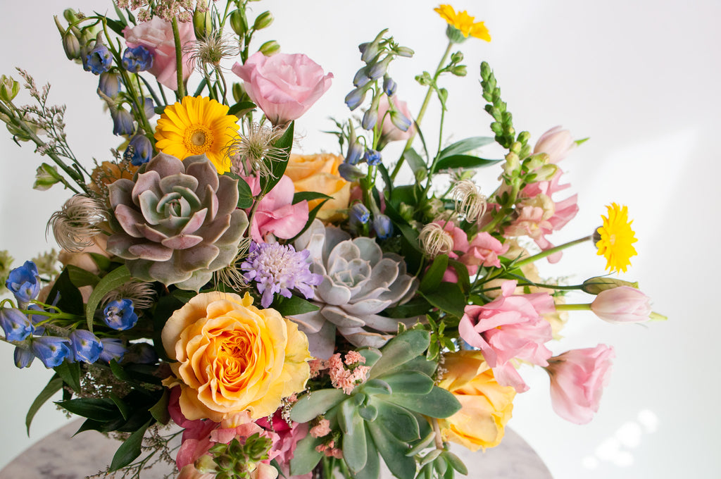 A flower arrangement in a glass vase in front of a white backdrop on a small marble table. It's filled with pastel pink, yellow, green, blue and purple flowers, including succulents, rose, delphinium, piccolini daisy, lisianthus, scabiosa, Queen Anne's lace, and tulip.