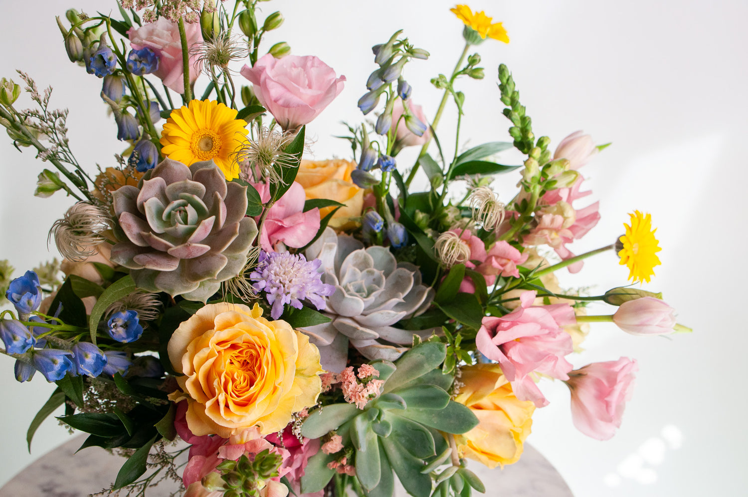 A flower arrangement in a glass vase in front of a white backdrop on a small marble table. It's filled with pastel pink, yellow, green, blue and purple flowers, including succulents, rose, delphinium, piccolini daisy, lisianthus, scabiosa, Queen Anne's lace, and tulip.