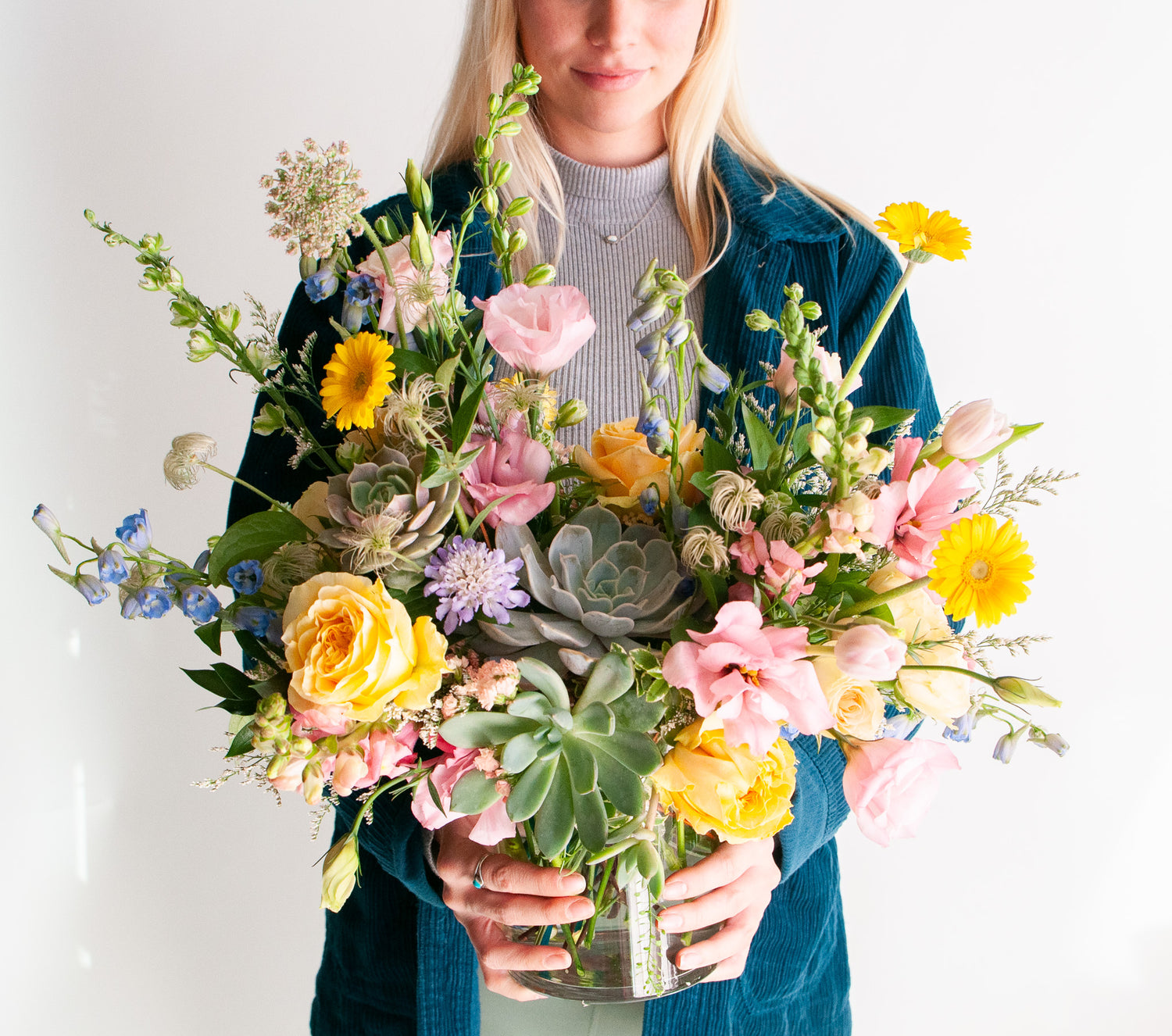 A woman holds a flower arrangement in a glass vase in front of a white backdrop. It's filled with pastel pink, yellow, green, blue and purple flowers, including succulents, rose, delphinium, piccolini daisy, lisianthus, scabiosa, Queen Anne's lace, and tulip.