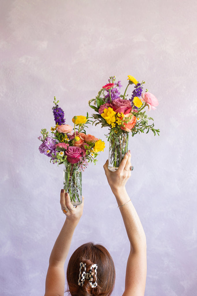 Woman holding WildFlora's Cheers to Mom Tumbler Pair in glass botanical tumblers in front of a light lilac backdrop. They have purple, pink, orange, & yellow flowers, including delphinium, piccolini daisy, daffodil, rose, ranunculus & chamomile.