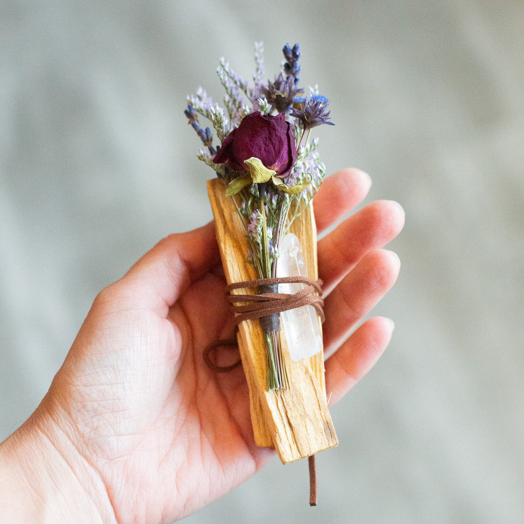 Hand holding a small bundle of dried palo santo, crystal, and flowers wrapped in twine against a neutral background