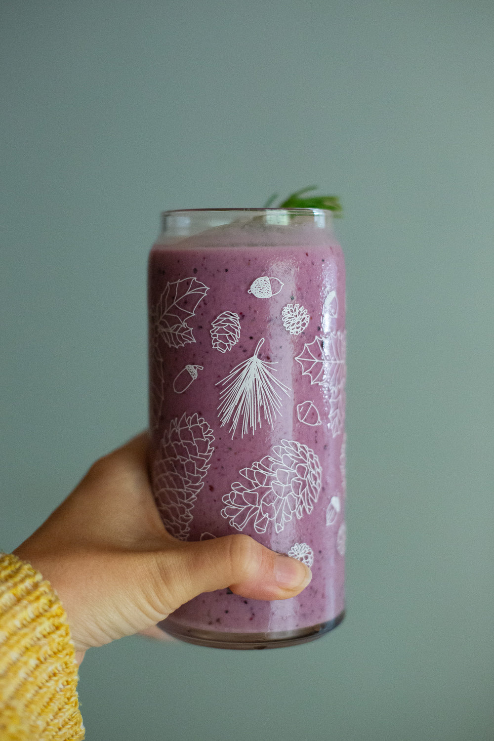 A hand holds a clear 16 ounce tumbler with a white conifer and pine cone graphic design. The tumbler is beer-can shaped and filled with a purple pink smoothie.