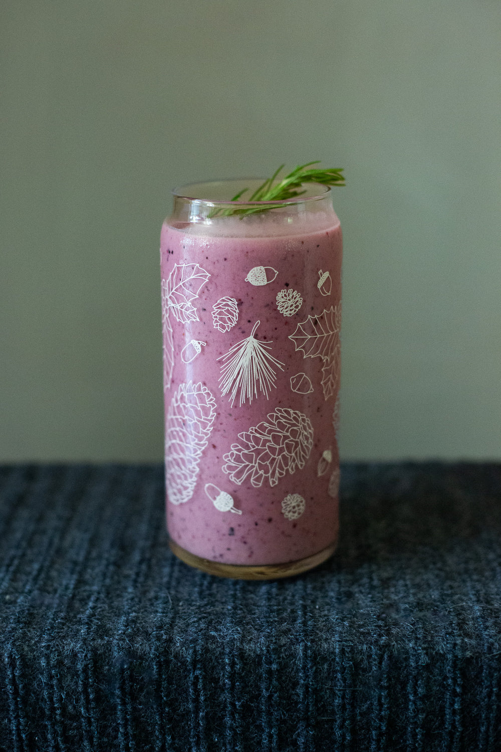 A clear 16 ounce tumbler with a white conifer and pine cone graphic design. The tumbler is beer-can shaped and filled with a purple pink smoothie.