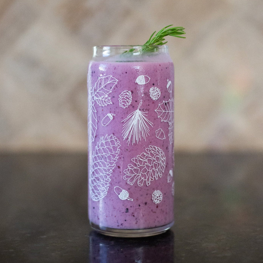 A clear 16 ounce tumbler with a white conifer and pine cone graphic design. The tumbler is beer-can shaped and filled with a purple pink smoothie.