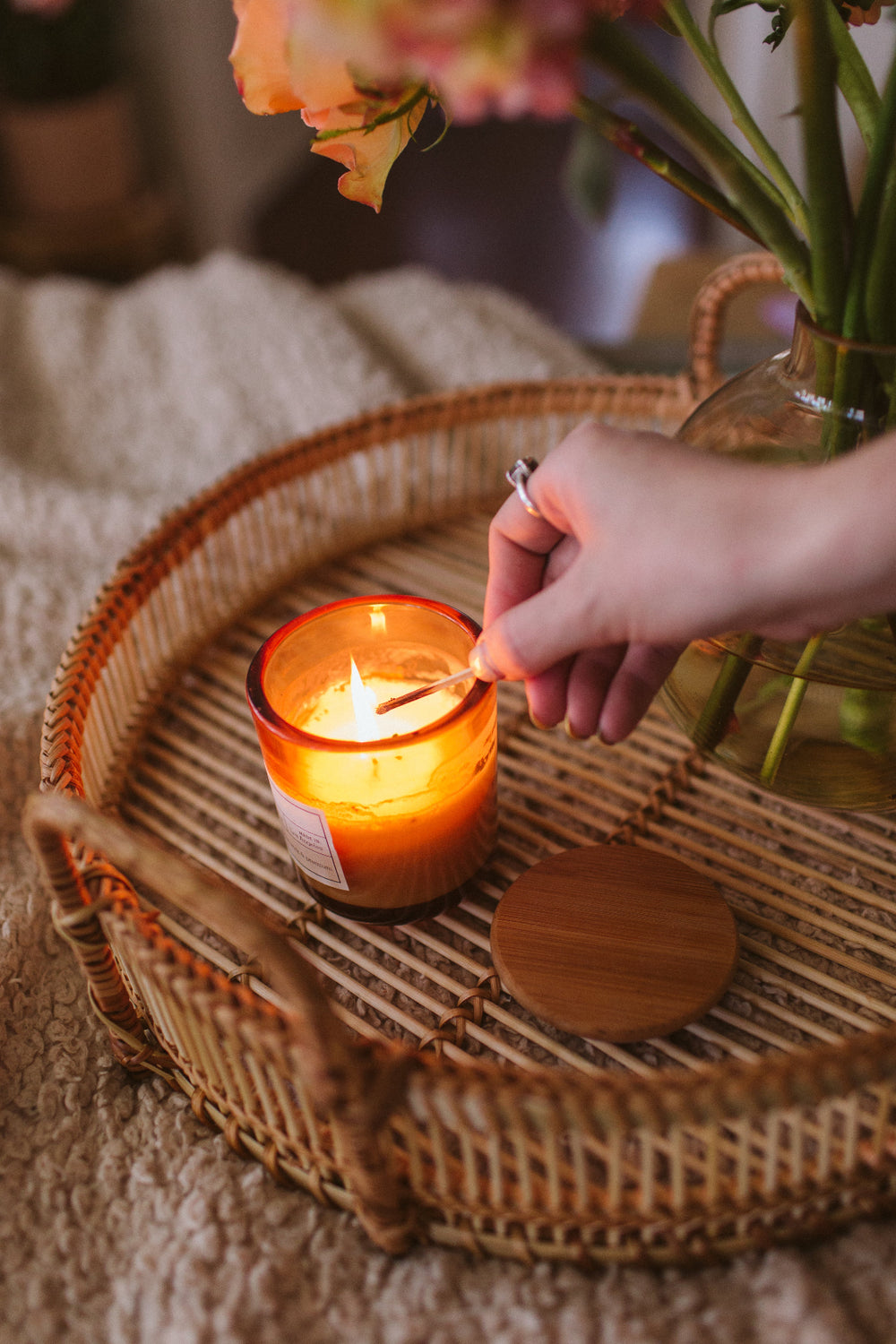 Person lighting a WildFlora single wick candle on a wicker tray with flowers in the background