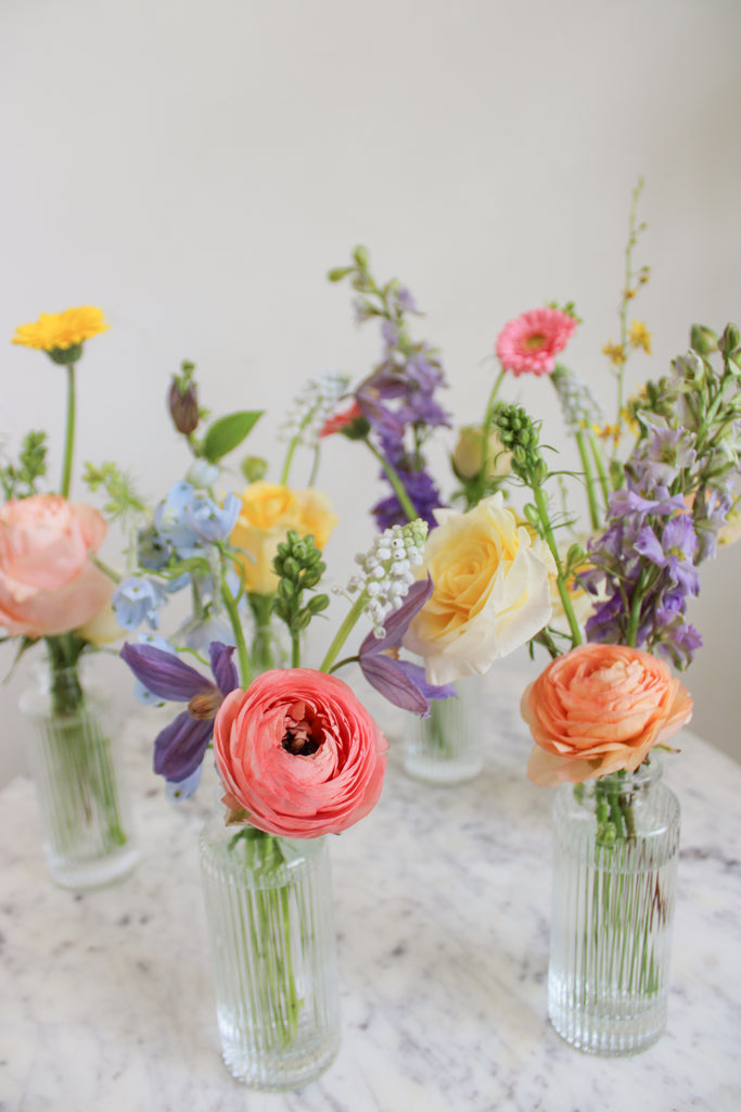 closeup of A set of clear glass bud vases with on a marble table in front of a white backdrop. They're filled with pink, peach, yellow blue and light purple flowers, including rose, piccolini daisy, clematis, ranunculus, liriope, and delphinium.