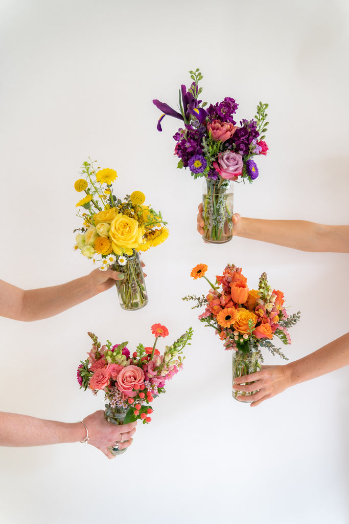 A set of 4 botanically-designed, beer-can shaped tumblers held in front of a white backdrop. One glass has pink flowers, the next orange, the next yellow, and the last purple, including rose, piccolini daisy, iris, billy ball, chamomile, tulip, snapdragon, and straw flowers blooms