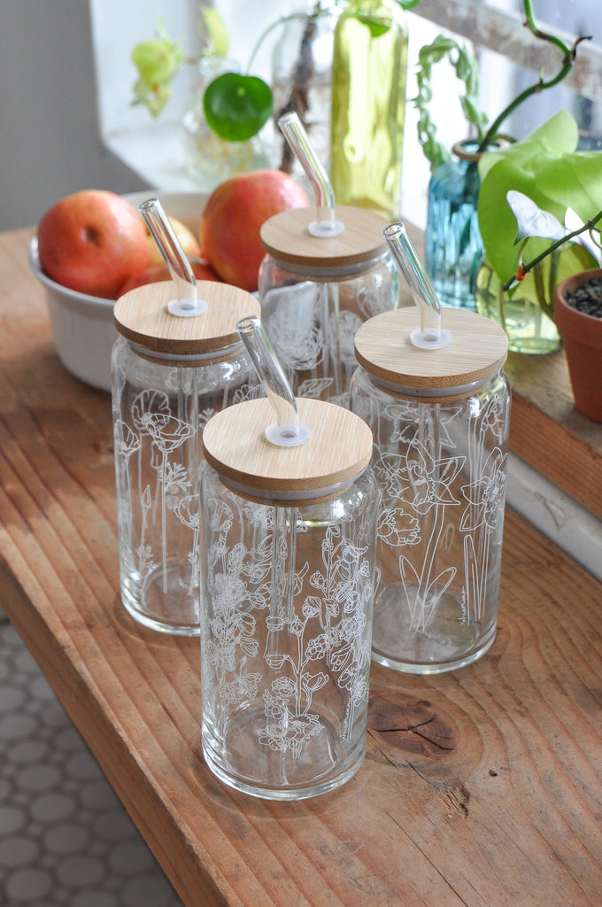 A set of 4 botanically-designed, beer-can shaped tumblers on a wood table, each with a wood lid and wide glass straw. They are screen printed with outlined drawings of flowers. behind is a window and a bowl of apples.