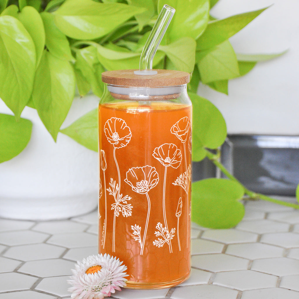 A clear 16 ounce tumbler with a white california poppy graphic design. The tumbler is beer-can shaped, has a wood lid with a wide glass straw, is adorned by a flower, and filled with an orange drink.