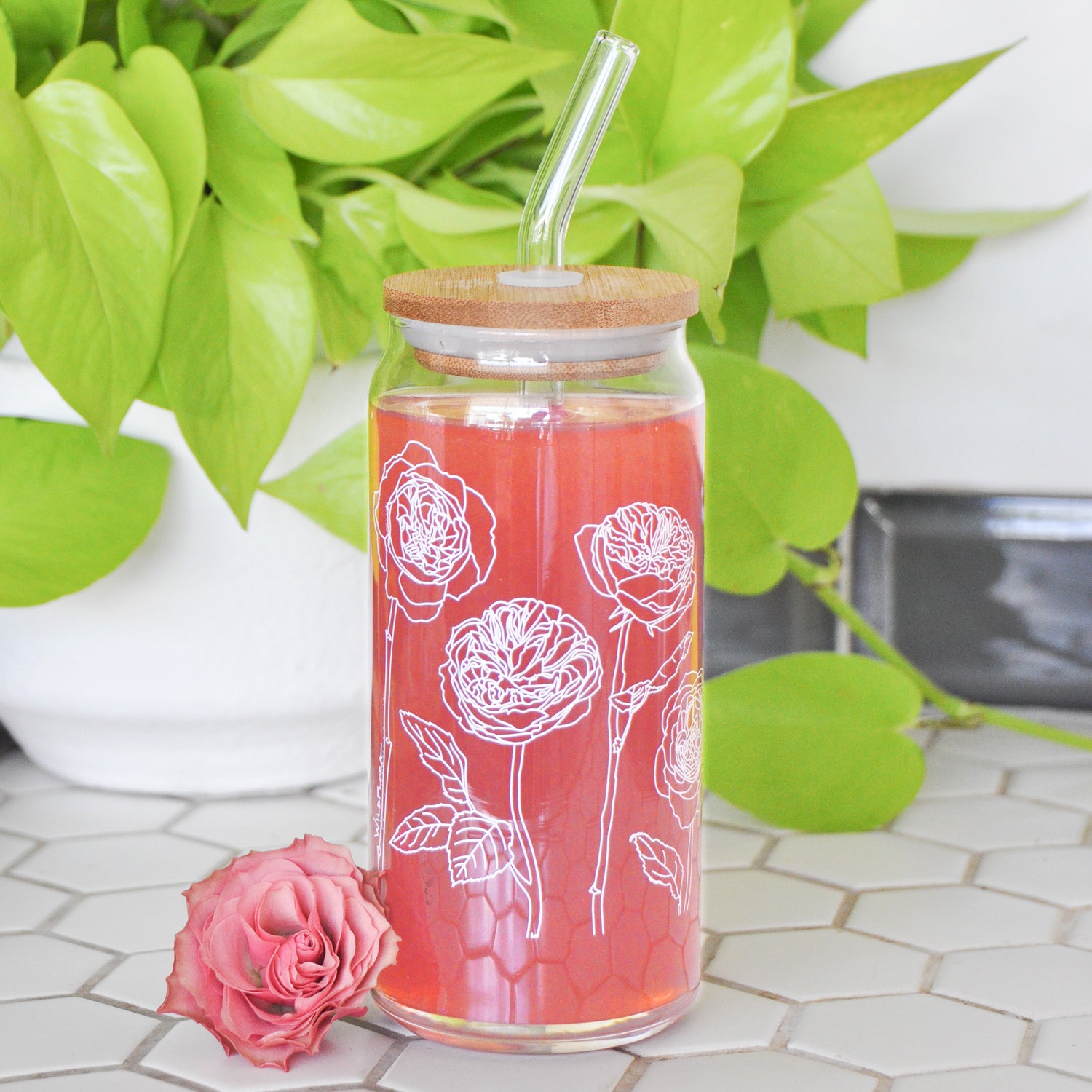 A clear 16 ounce tumbler with a white garden rose graphic design. The tumbler is beer-can shaped, has a wood lid with a wide glass straw, is adorned by a flower, and filled with a pink drink.