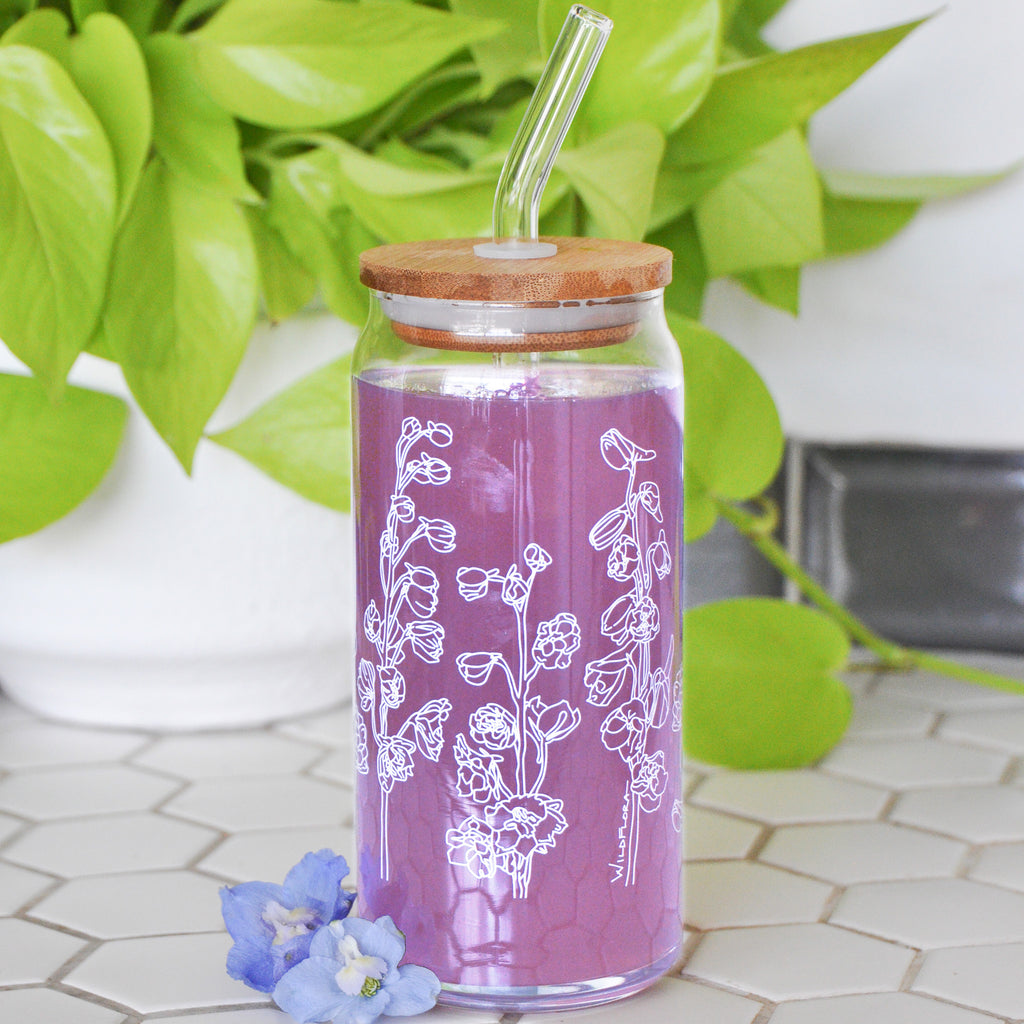 A clear 16 ounce tumbler with a white delphinium graphic design. The tumbler is beer-can shaped, has a wood lid with a wide glass straw, is adorned by a flower, and filled with a purple drink.