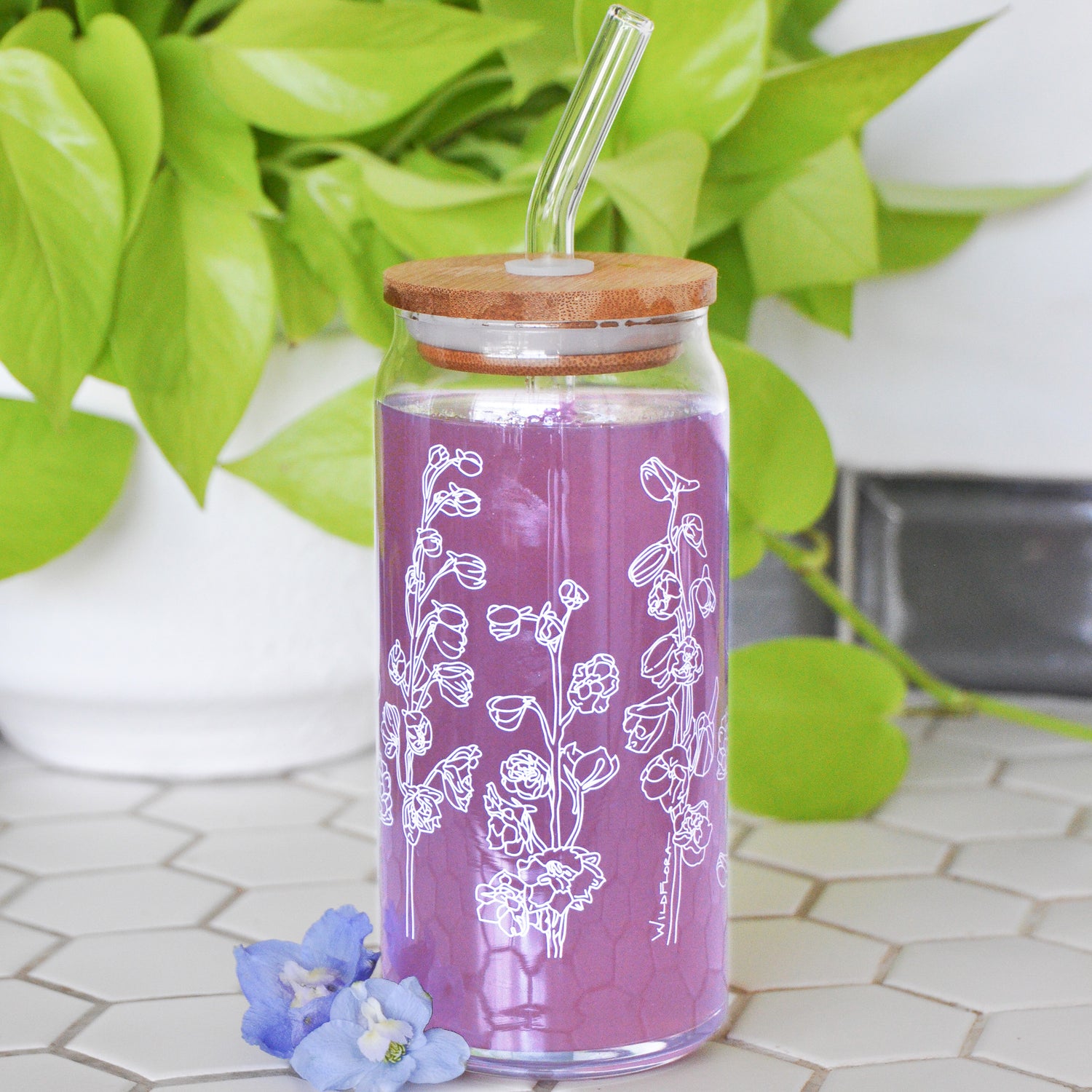 A clear 16 ounce tumbler with a white delphinium graphic design. The tumbler is beer-can shaped, has a wood lid with a wide glass straw, is adorned by a flower, and filled with a purple drink.