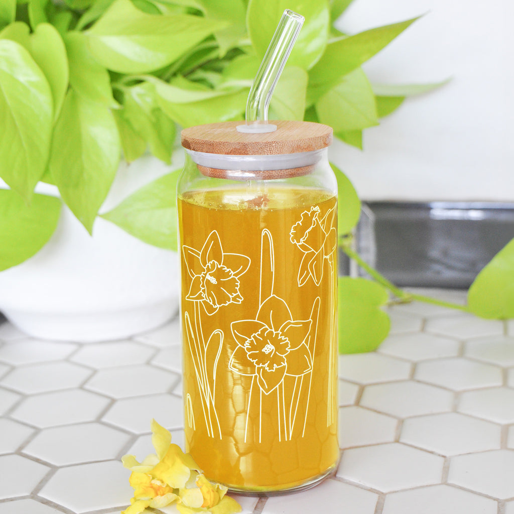 A clear 16 ounce tumbler with a white daffodil graphic design. The tumbler is beer-can shaped, has a wood lid with a wide glass straw, is adorned by a flower, and filled with a yellow drink.