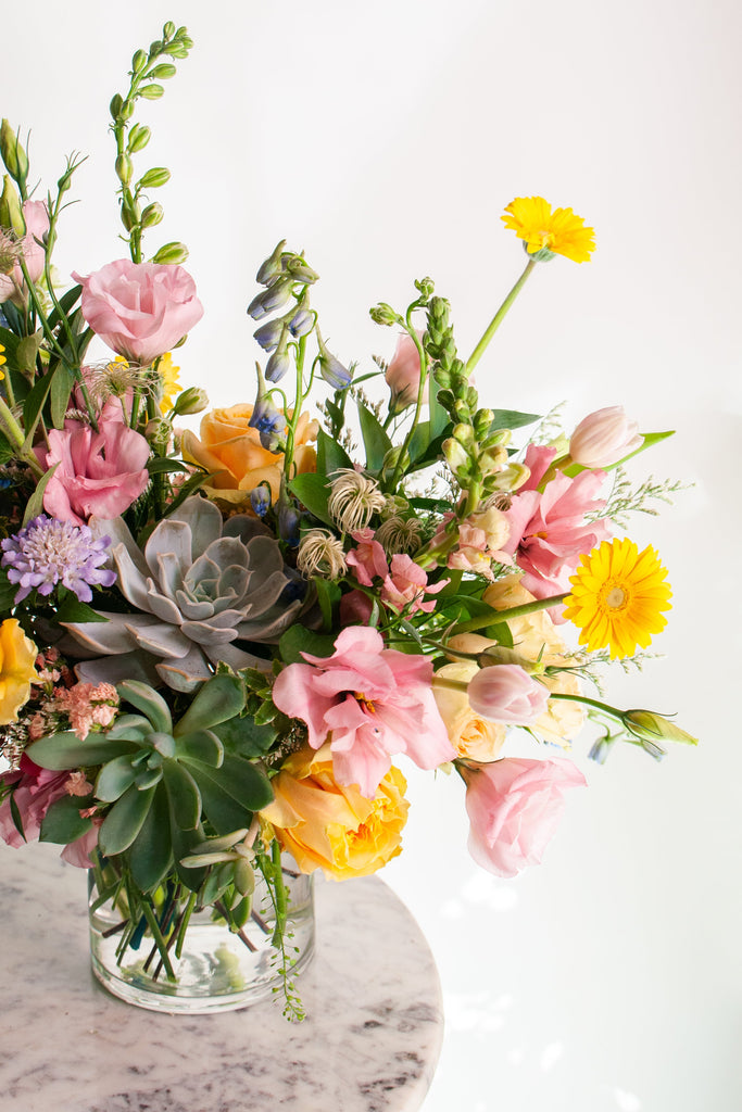 A flower arrangement in a glass vase in front of a white backdrop on a small marble table. It's filled with pastel pink, yellow, green, blue and purple flowers, including succulents, rose, delphinium, piccolini daisy, lisianthus, scabiosa, Queen Anne's lace, and tulip.