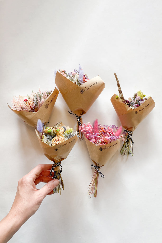 A flat lay of mini dried floral bouquets about the size of a hand. they are in varying colors and wrapped in brown kraft paper that is stamped with a WildFlora logo.