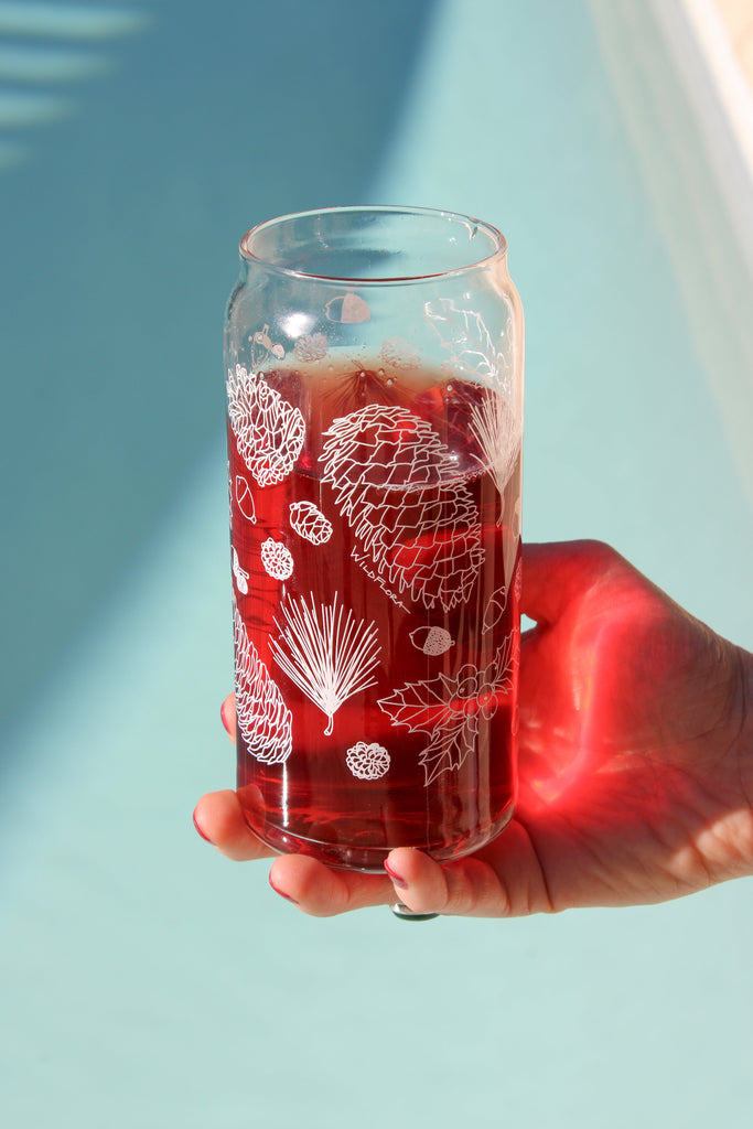 A hand holds a clear 16 ounce tumbler with a white conifer and pine cone graphic design. The tumbler is beer-can shaped and filled with a red drink.