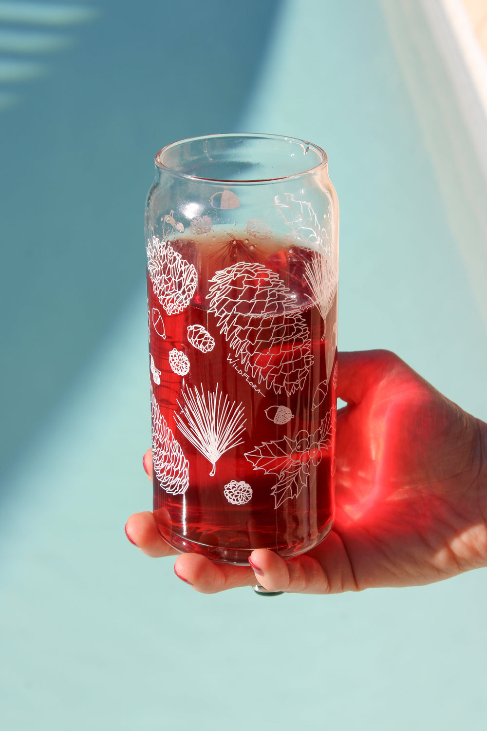 A hand holds a clear 16 ounce tumbler with a white conifer and pine cone graphic design. The tumbler is beer-can shaped and filled with a red drink.