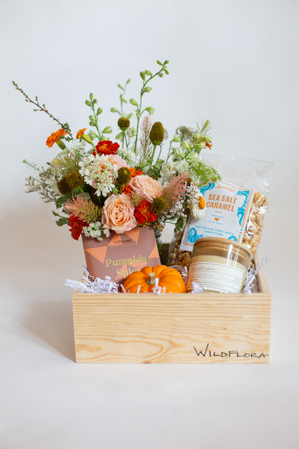 A Wildflora gift box with peach, orange, red, and white flowers, a small pumpkin, a candle, and sea salt popcorn in a wood box.