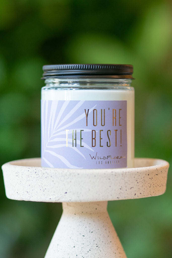 Expressions candle that says "you're the best!" sits on a candle holder in front of greens. Single-wick, white soy wax candle in a glass container with a black lid.