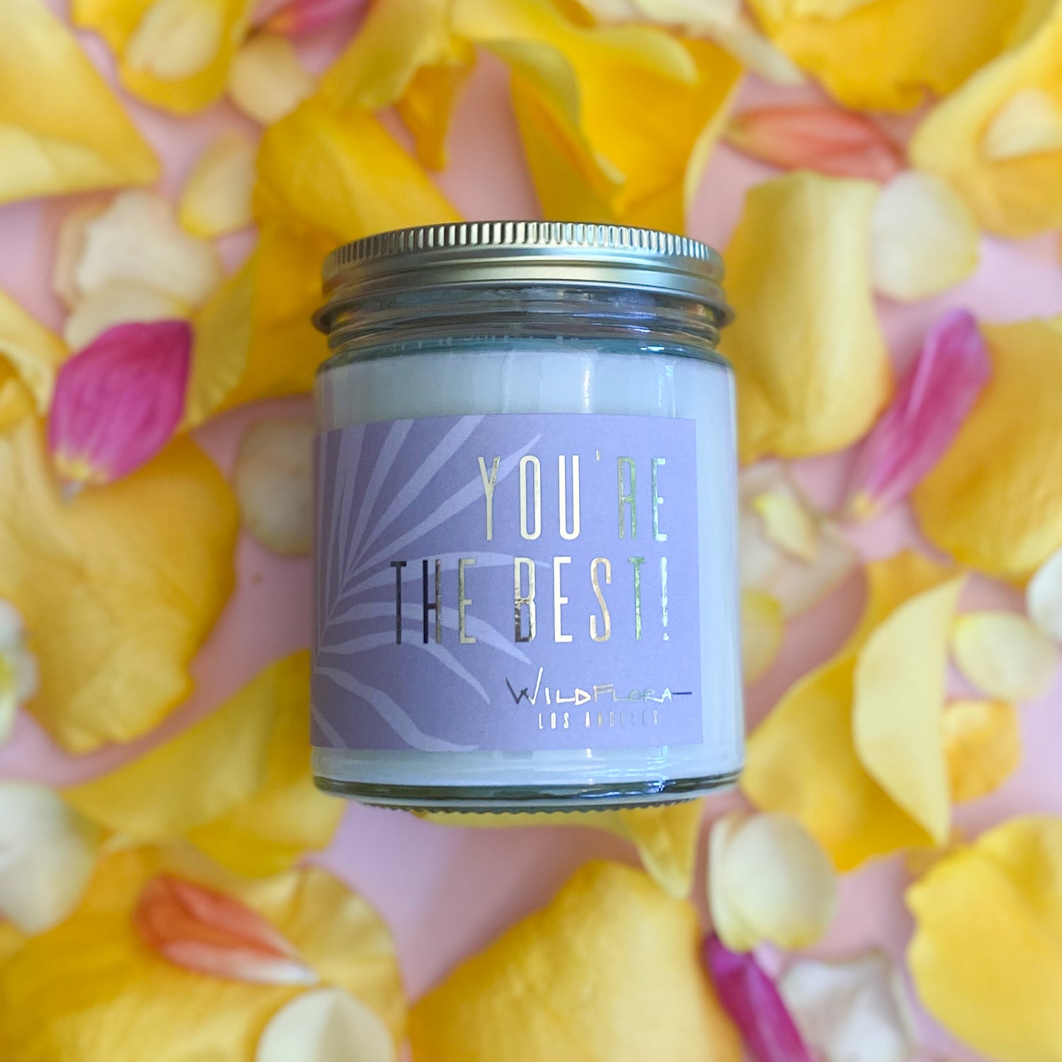 Expressions candle that says "you're the best!" lays on a pick background with yellow and pink petals. Single-wick, white soy wax candle in a glass container with a black lid.