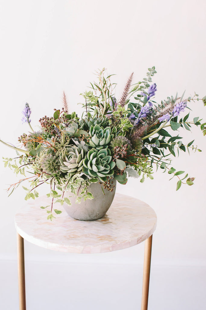 A flower arrangement in a concrete container in front of a white backdrop on a small table. It's filled with green and purple-colored flowers and foliage, including succulents, eucalyptus, ornamental grass, and hyacinth. It's got a wild, natural vibe to it.