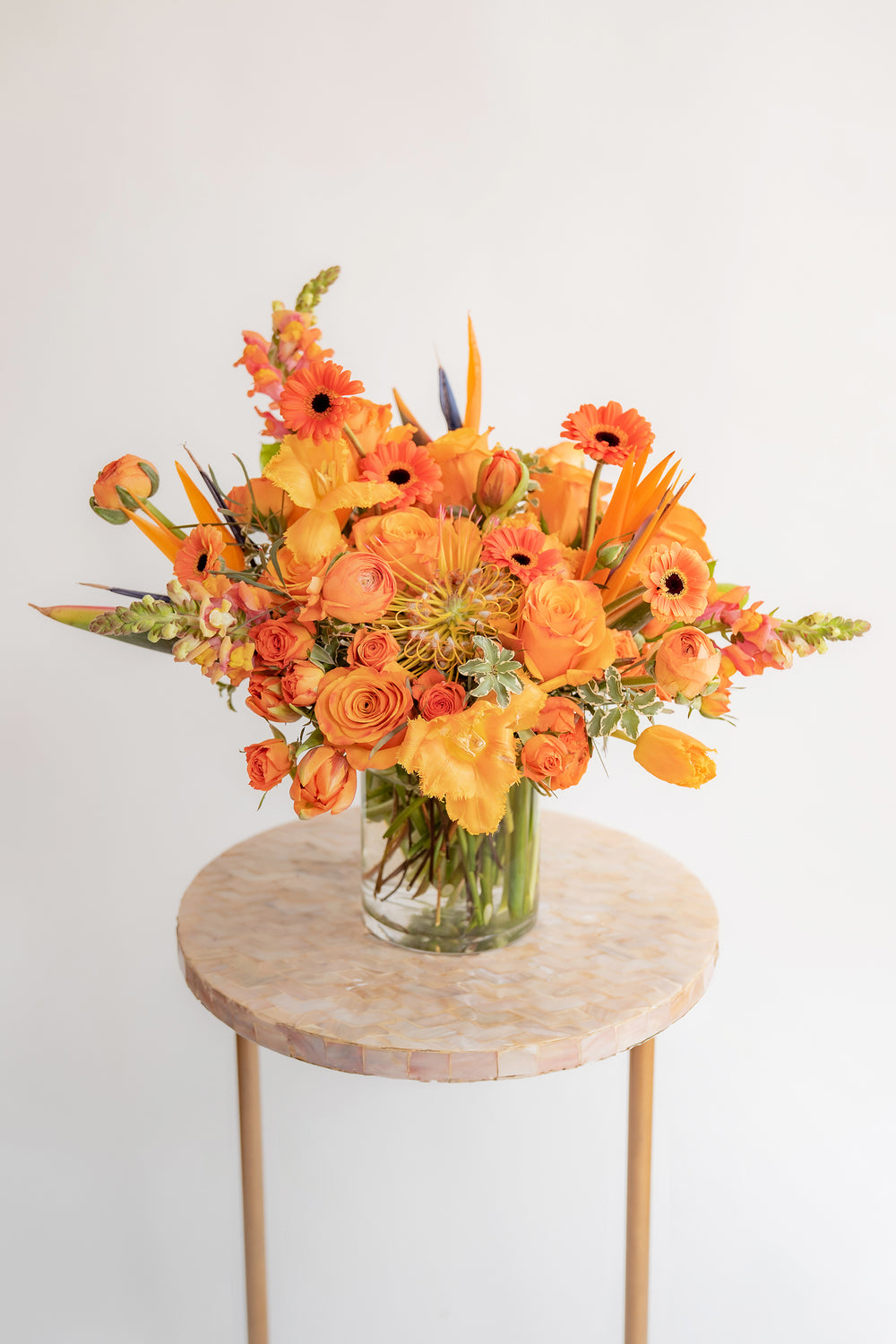 A flower arrangement in a glass container in front of a white backdrop on a small table. It's filled with orange and tangerine-colored flowers, including rose, snapdragon, piccolini daisy, bird of paradise, tulips, and pincushion protea.