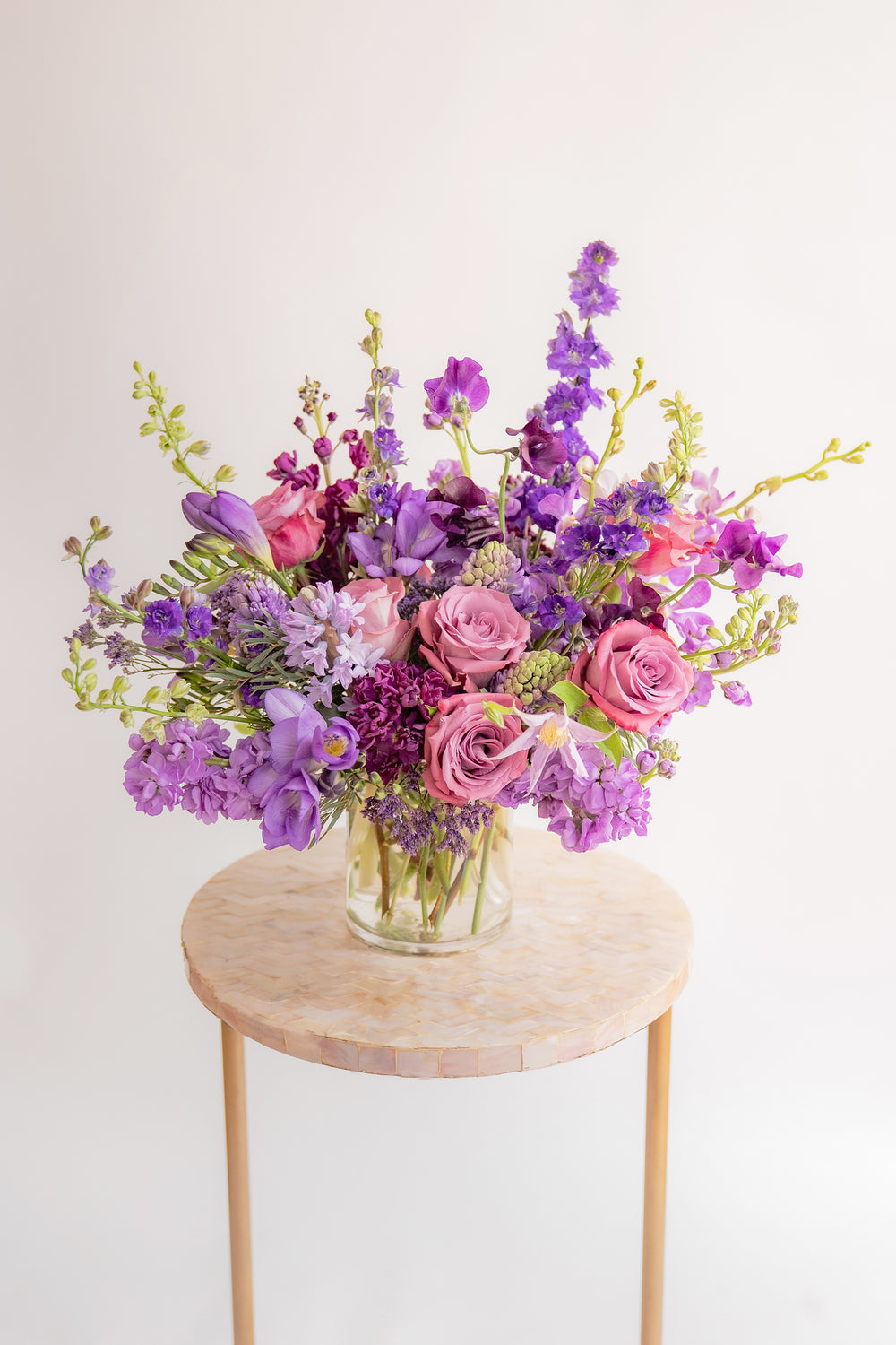 A flower arrangement in a glass container in front of a white backdrop on a small table. It's filled with lavender and purple flowers, including rose, stock, delphinium, and statice, hyacinth, and clematis.