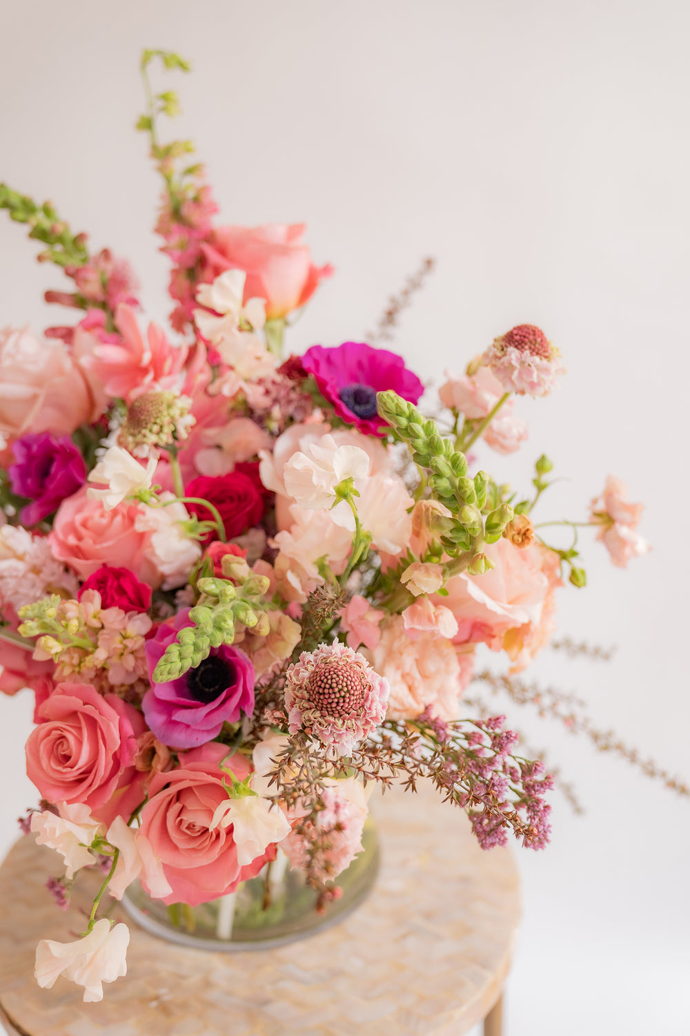 closeup of A flower arrangement in a glass container in front of a white backdrop on a small table. It's filled with pink and magenta flowers, including rose, scabiosa, sweet pea, snapdragon, and delphinium.