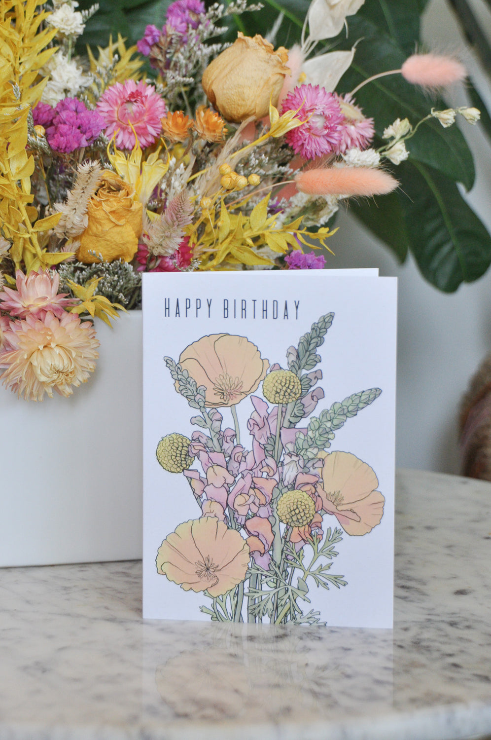 A greeting card that says "happy birthday" and has artwork of california poppies, billy balls, and snapdragon on it. It sits on a marble table and behind are dried flowers.