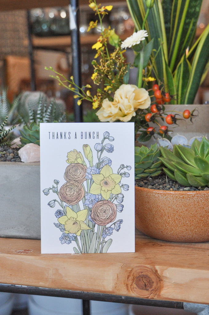 A greeting card that says "thanks a bunch" and has artwork of ranunculus, daffodils, and delphinium on it. It sits on a wood table and behind it are potted plants.