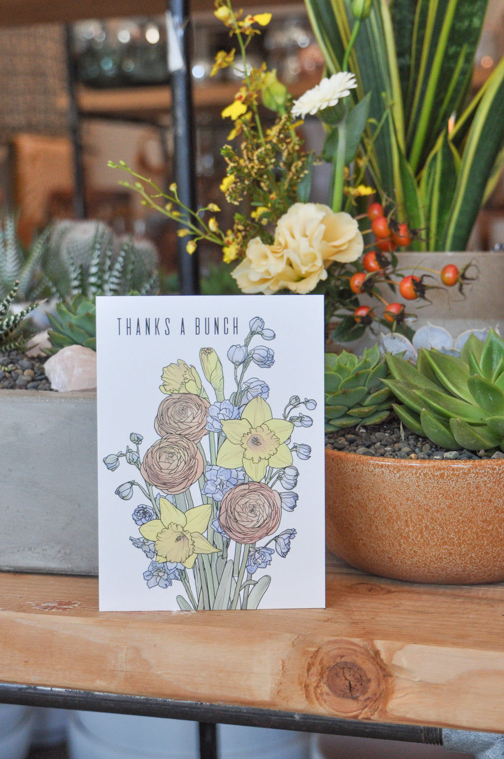 A greeting card that says "thanks a bunch" and has artwork of ranunculus, daffodils, and delphinium on it. It sits on a wood table and behind it are potted plants.