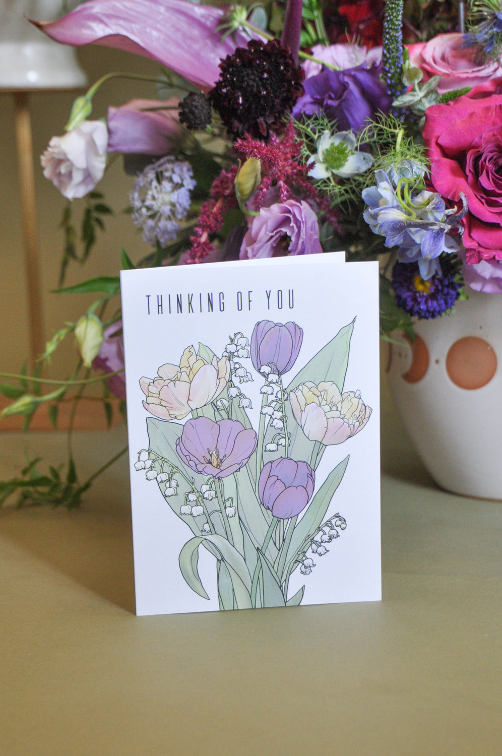 A greeting card that says "thinking of you" and has artwork of tulips and lily of the valley on it. It's standing on a table and behind are purple flowers.