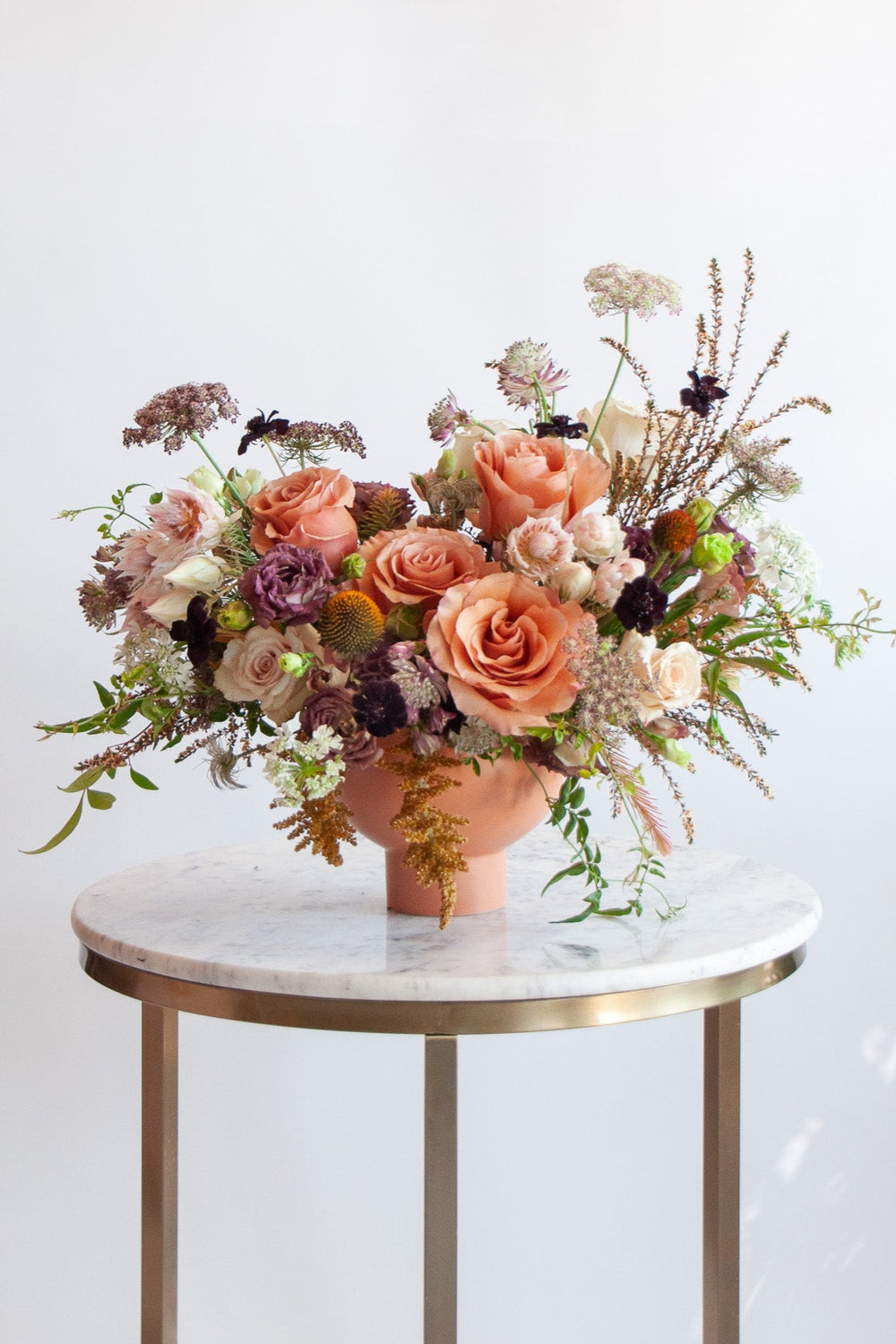 A cream, eggplant, salmon and peach WildFlora flower arrangement in a terracotta compote vase, featuring Moab rose, thistle, Queen Anne's lace, blushing bride protea, and lisianthus.