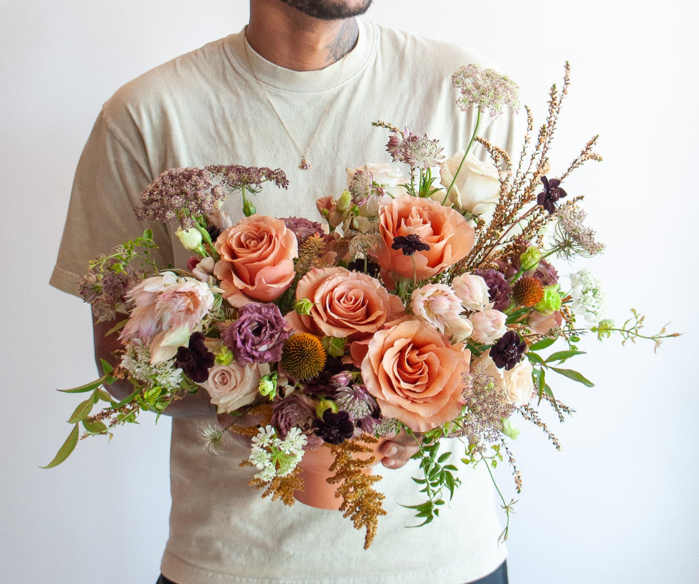 Man holding a cream, eggplant, salmon and peach WildFlora flower arrangement in a terracotta compote vase, featuring Moab rose, thistle, Queen Anne's lace, blushing bride protea, and lisianthus.