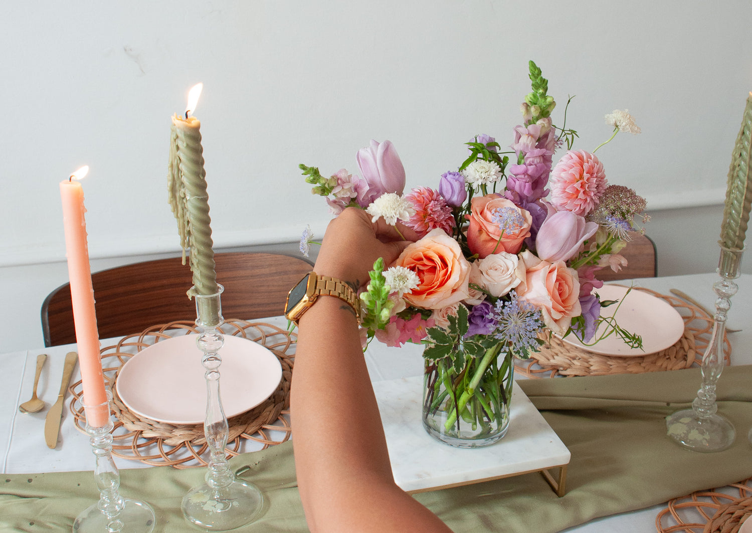 A orange, peach, pink, purple, lavender, chartreuse and white flower centerpiece sits on an event table with pink plate, gold cutlery, woven plate chargers, and tall candlesticks. The floral arrangement includes roses, dahlias, clematis, allium, snapdragon, queen anne's lace, and lisianthus. The vibe is sweet, romantic, and dreamy.