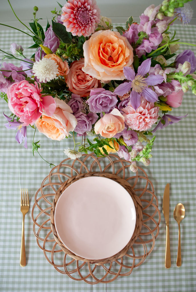 A orange, peach, pink, purple, lavender, chartreuse and white flower centerpiece sits on an event table with pink plate, gold cutlery, woven plate chargers, and tall candlesticks. The floral arrangement include, roses, dahlias, peonies, clematis, allium, snapdragon, amaranthus, and lisianthus. The vibe is sweet, romantic, and dreamy.