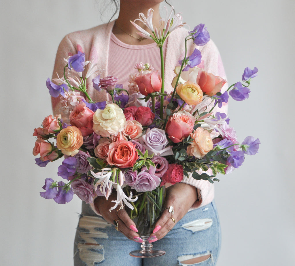 A woman holds a flower arrangement in a glass vase in front of a white backdrop. It's filled with pink, coral, purple, peach, orange, and violet-colored flowers, including rose, ranunculus, sweet pea, lily, and stock.