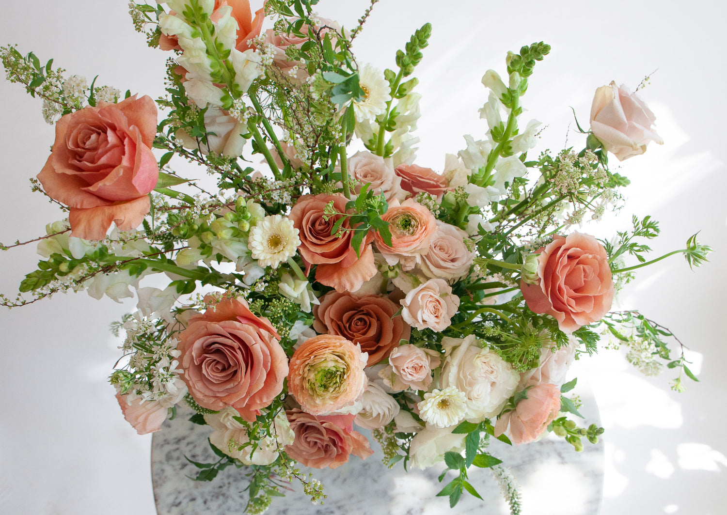 Close-up of A flower arrangement in a glass vase on a marble table with green, peach, white, and salmon orange-colored flowers, including rose, piccolini daisy, ranunculus, snapdragon, and Queen Anne's lace. The backdrop is white.