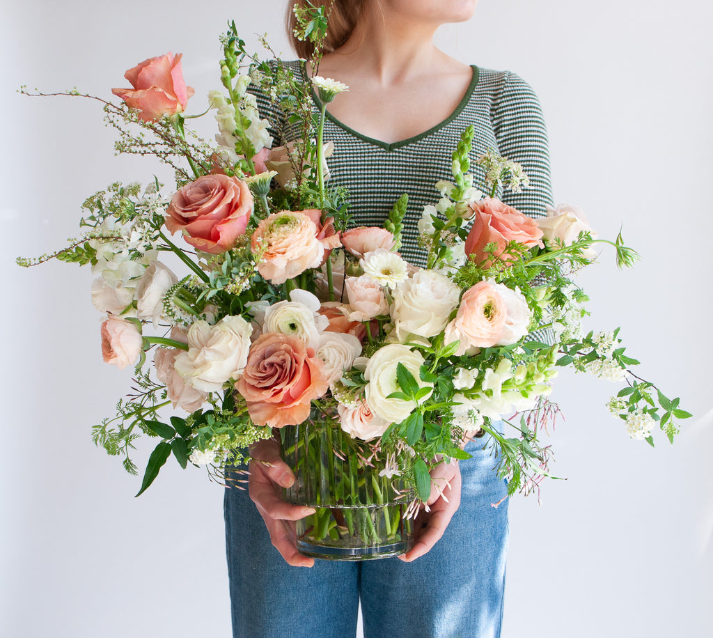 A woman holding A flower arrangement in a glass vase with green, peach, white, and salmon orange-colored flowers, including rose, piccolini daisy, ranunculus, snapdragon, and Queen Anne's lace. The backdrop is white.