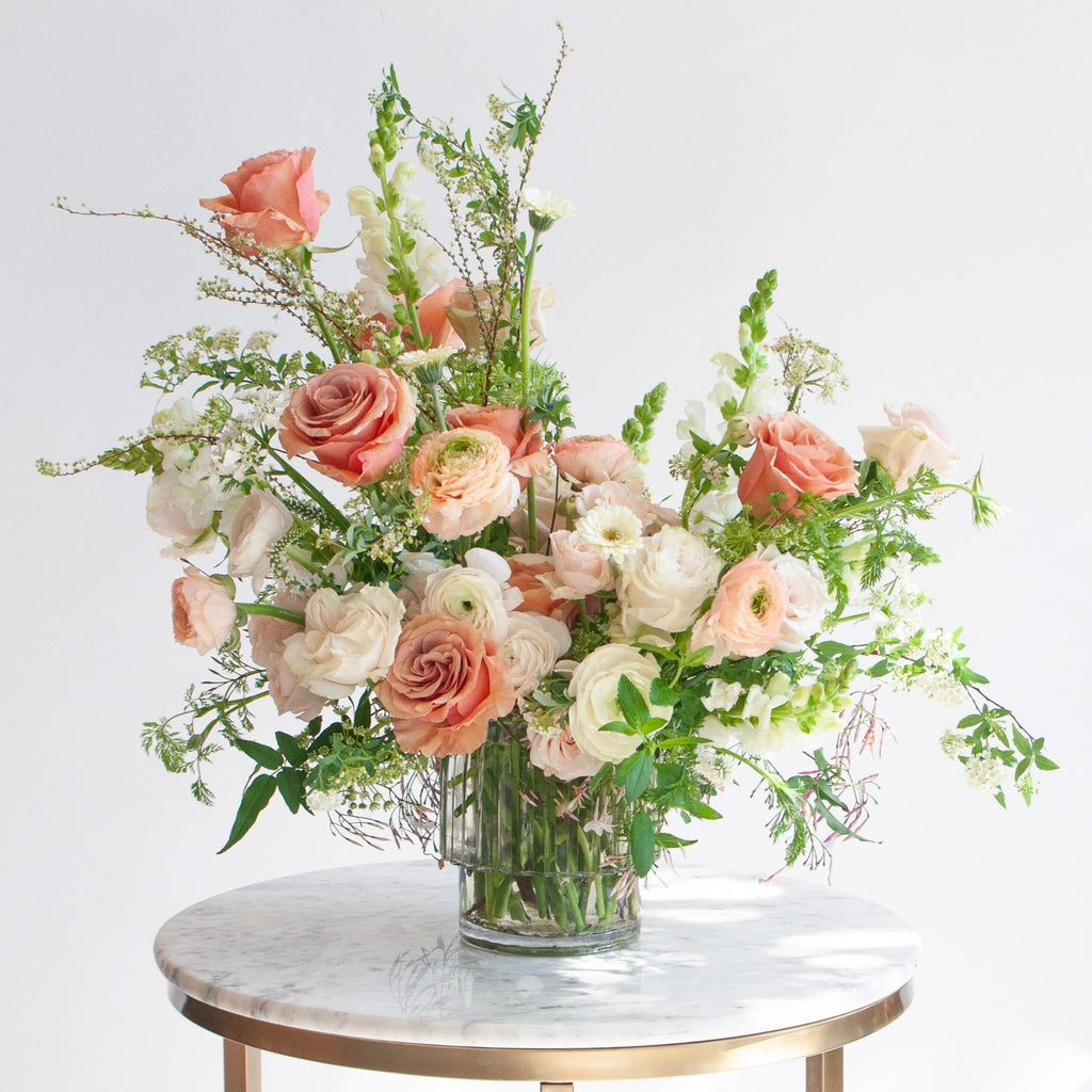 A flower arrangement in a glass vase on a marble table with green, peach, white, and salmon orange-colored flowers, including rose, piccolini daisy, ranunculus, snapdragon, and Queen Anne's lace. The backdrop is white.