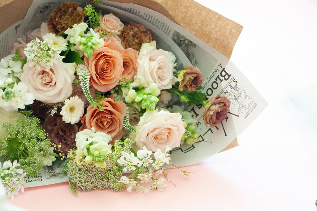 closeup of a bouquet wrapped in floral newspaper and brown kraft paper in front of a white backdrop. It's filled with peach, tan, white, and orange flowers, including rose, piccolini daisy, lisianthus, queen anne's lace, allium, and veronica.