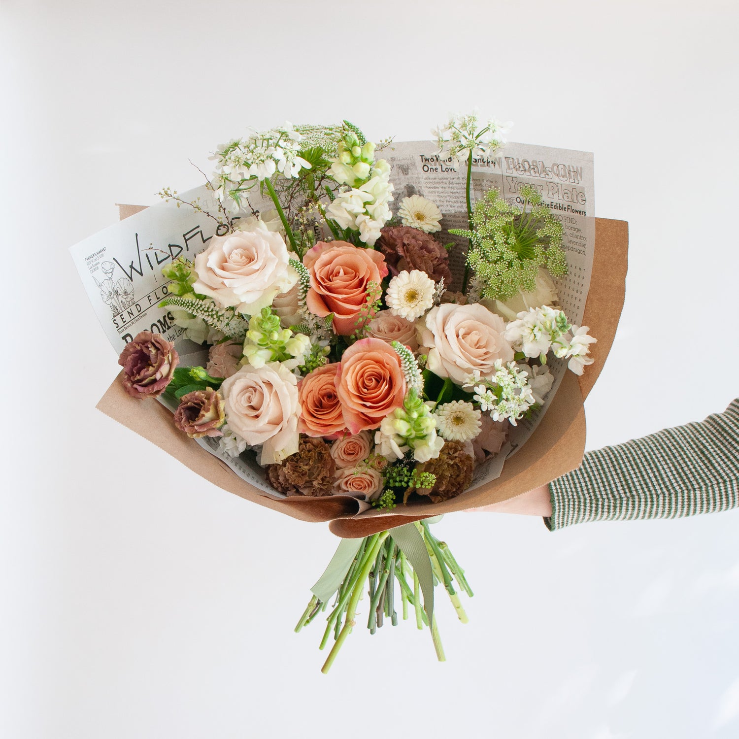 An arm holds a bouquet wrapped in floral newspaper and brown kraft paper in front of a white backdrop. It's filled with peach, tan, white, and orange flowers, including rose, piccolini daisy, lisianthus, queen anne's lace, allium, and veronica.