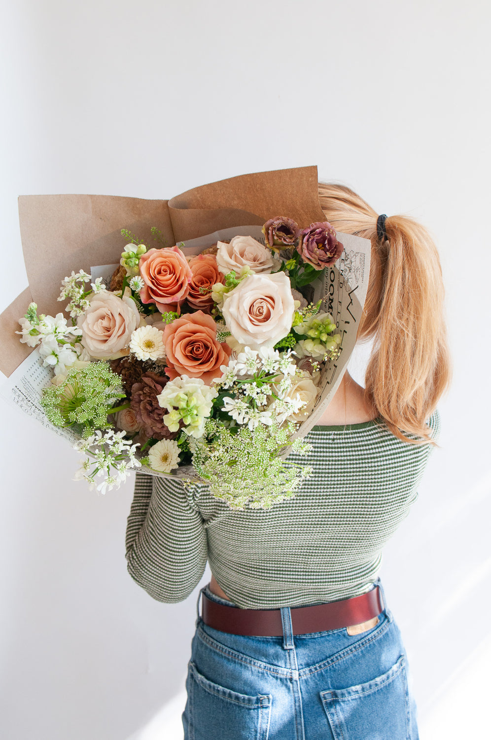 view from behind a woman who holds a bouquet wrapped in floral newspaper and brown kraft paper over her shoulder in front of a white backdrop. It's filled with peach, tan, white, and orange flowers, including rose, piccolini daisy, lisianthus, queen anne's lace, allium, and veronica.